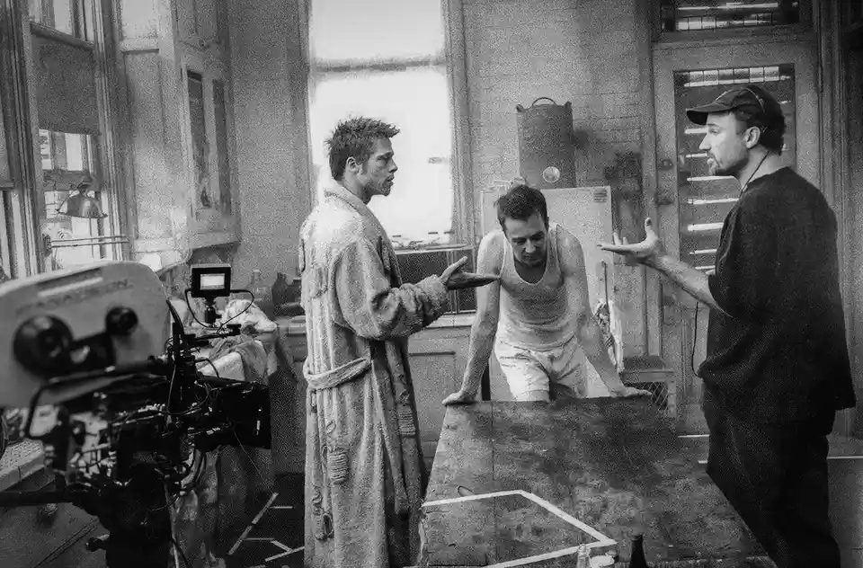 A black-and-white behind-the-scenes photo from a film set shows three men in a gritty room. Brad Pitt, in a tattered robe…