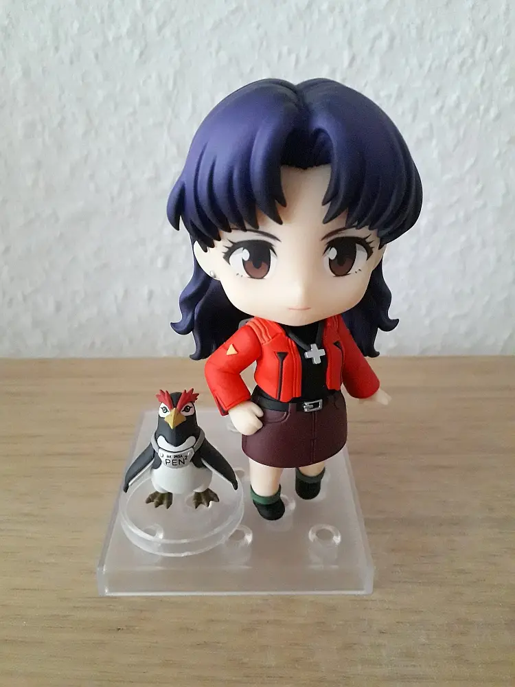 A chibi-style anime figurine of a girl with dark blue hair, wearing a red jacket and brown skirt, stands next to a small…