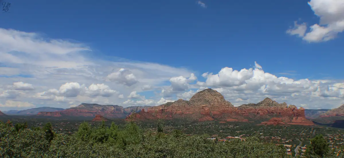 A scenic landscape photo of red rock formations under a blue sky with scattered white clouds, with green trees and a distant…