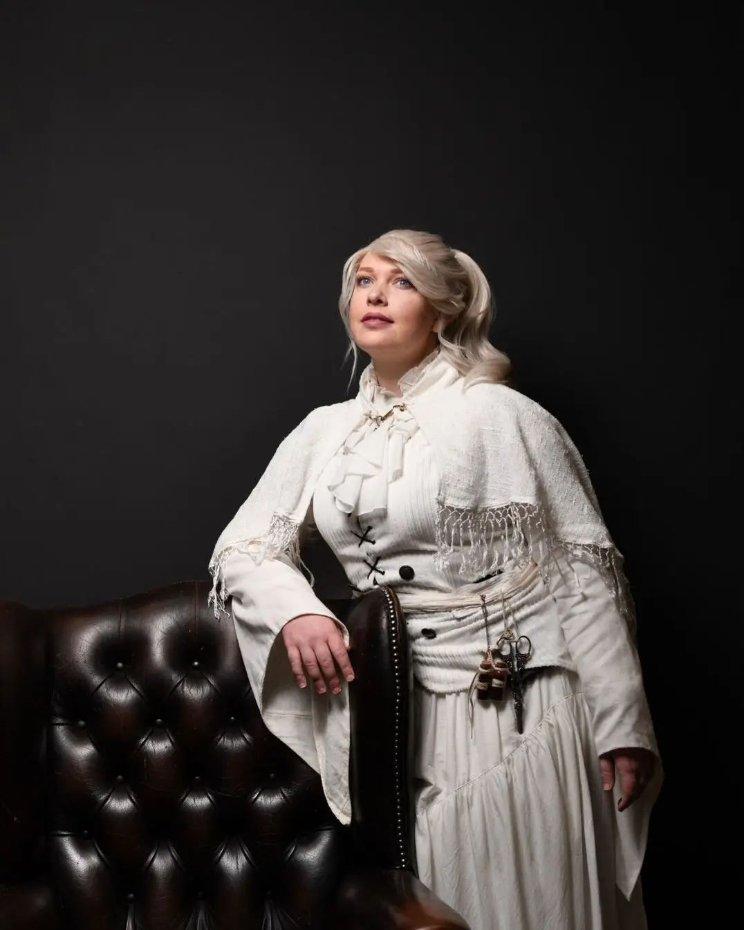 A woman with blonde hair in a white Victorian-style dress stands beside a dark brown leather tufted armchair against a black…