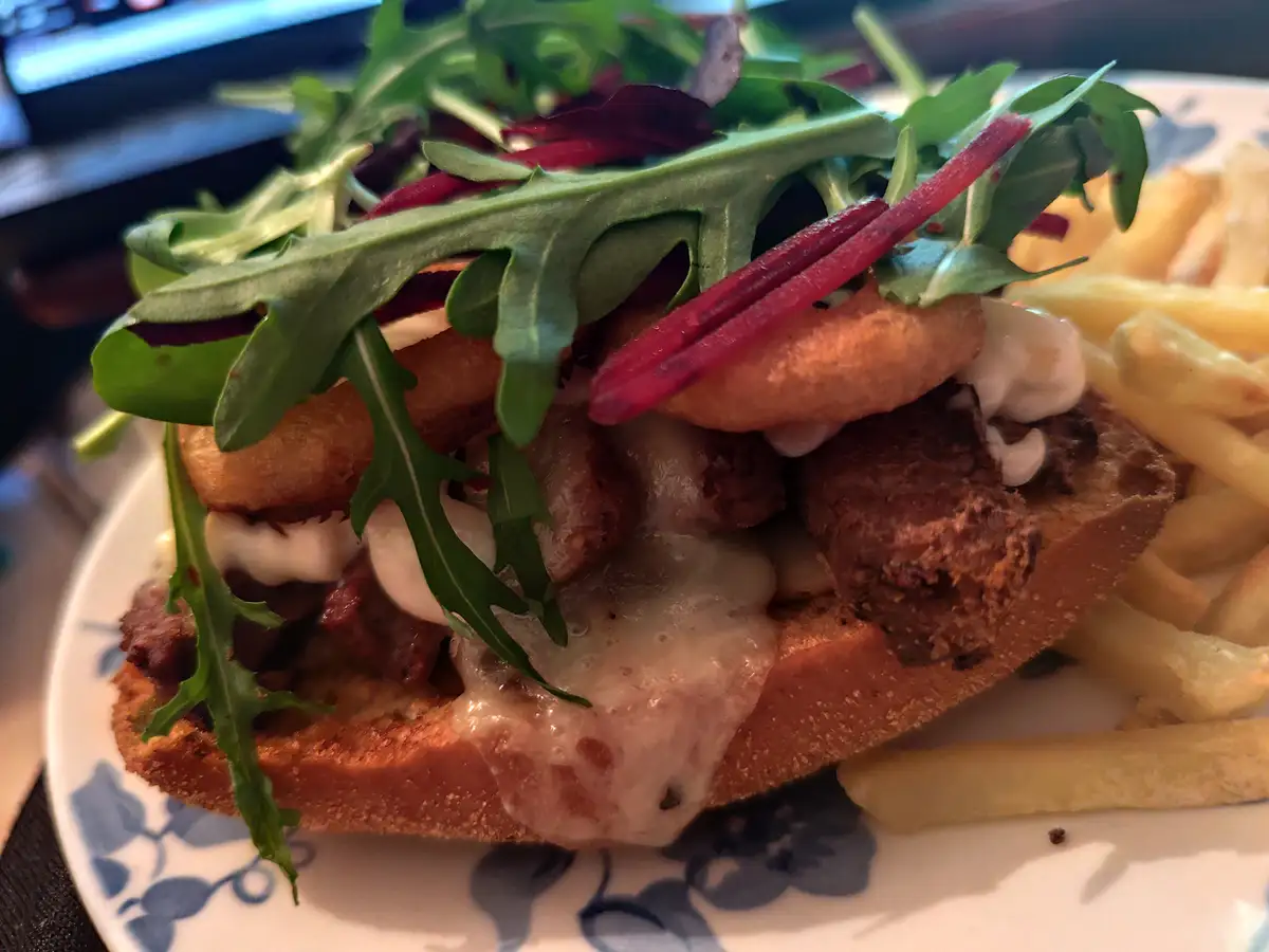 A close-up photo of a gourmet burger on a white plate with blue floral patterns, topped with arugula, red beet slices, fried…