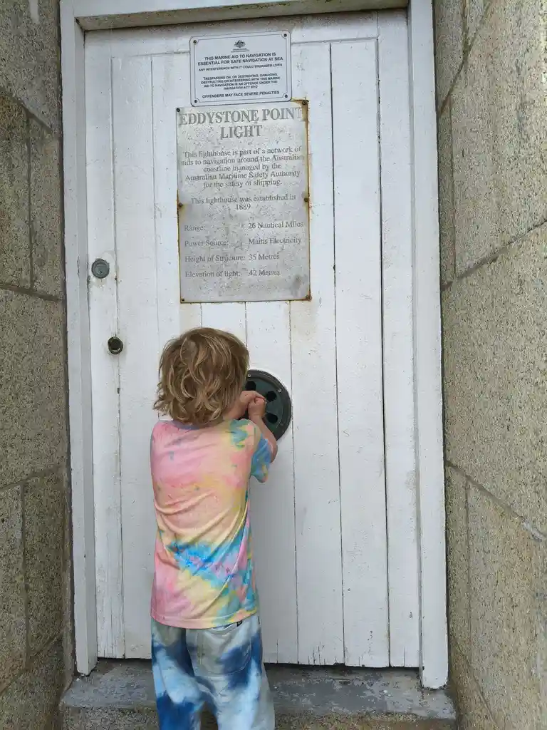 A young child in a tie-dye shirt peers through a small, circular porthole in a white wooden door of a lighthouse. Above the…