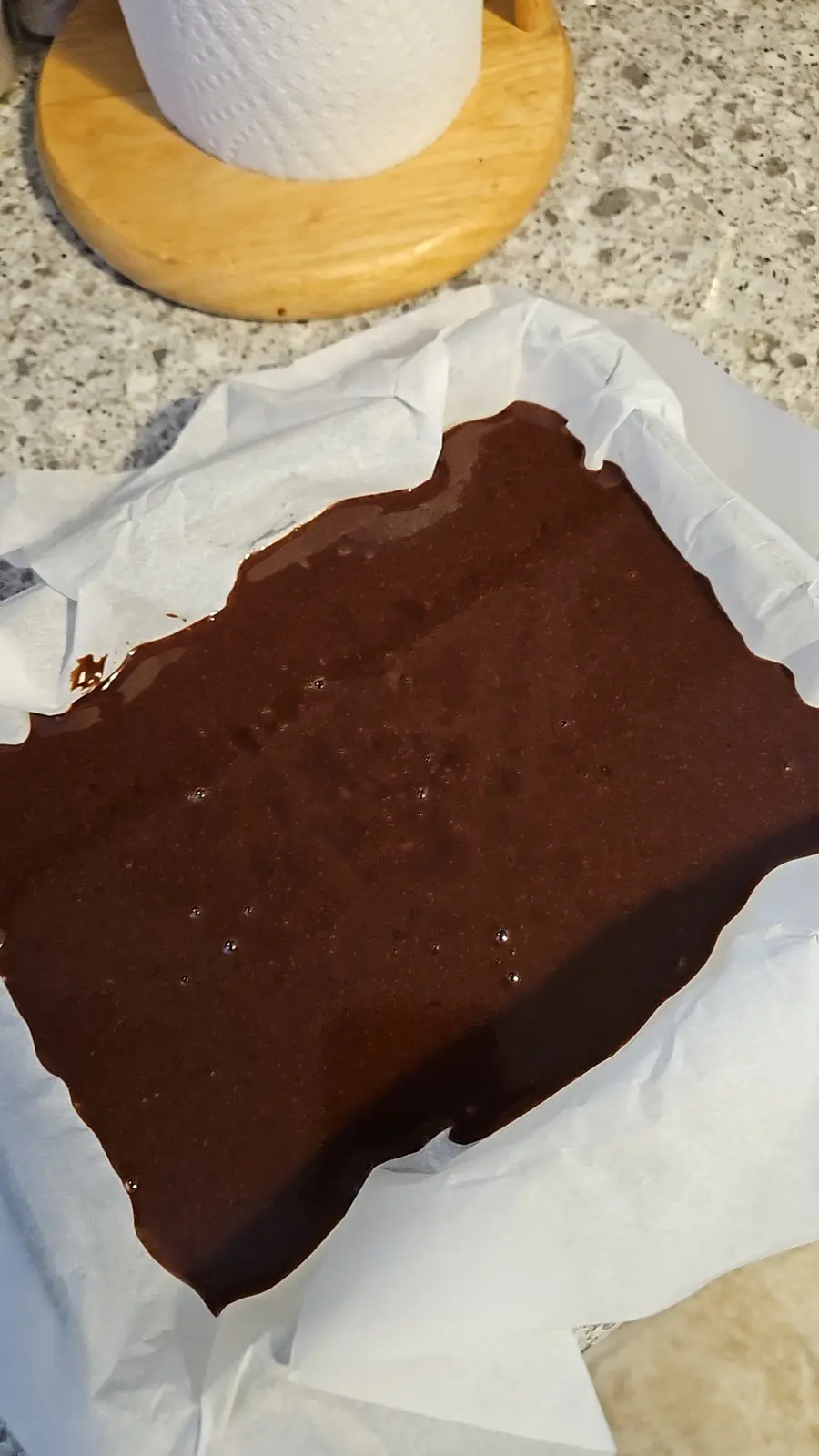 A close-up photo shows a dark brown, glossy chocolate cake batter that has overflowed from a baking pan lined with white…