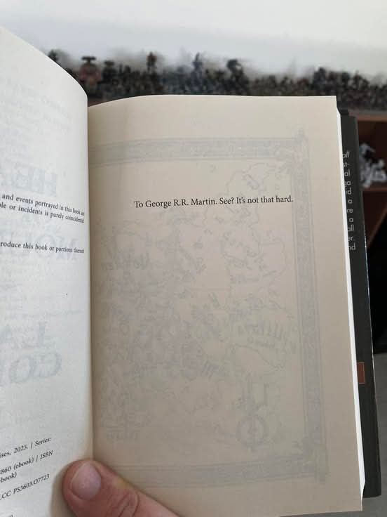 An open book shows a dedication page with the text 'To George R.R. Martin. See? It's not that hard.' The background behind…