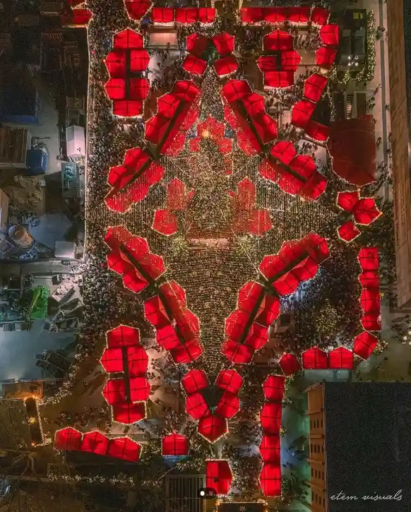 An aerial nighttime photo shows a crowded city square decorated with a large, glowing Christmas tree and numerous red…