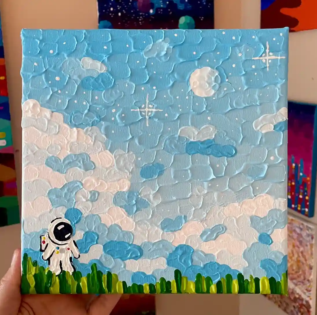 A hand holds a textured, thickly painted canvas depicting a small astronaut standing on green grass under a sky filled with…