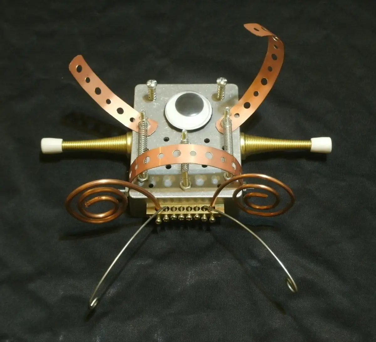 A handmade robot constructed from metal parts, including copper strips shaped like antennae and legs, brass rods with white…