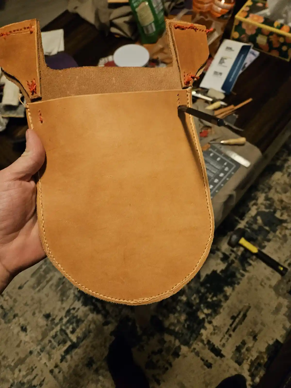 A hand holds up a handmade leather object shaped like a toilet seat, with visible stitching and a tool inserted into one…