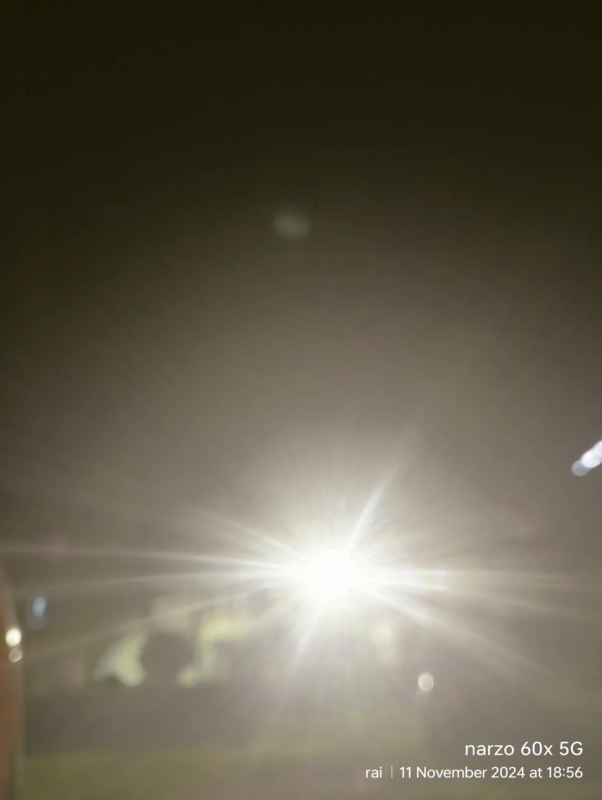 A blurry nighttime photo showing a bright, overexposed light source with lens flare radiating outward, obscuring most…