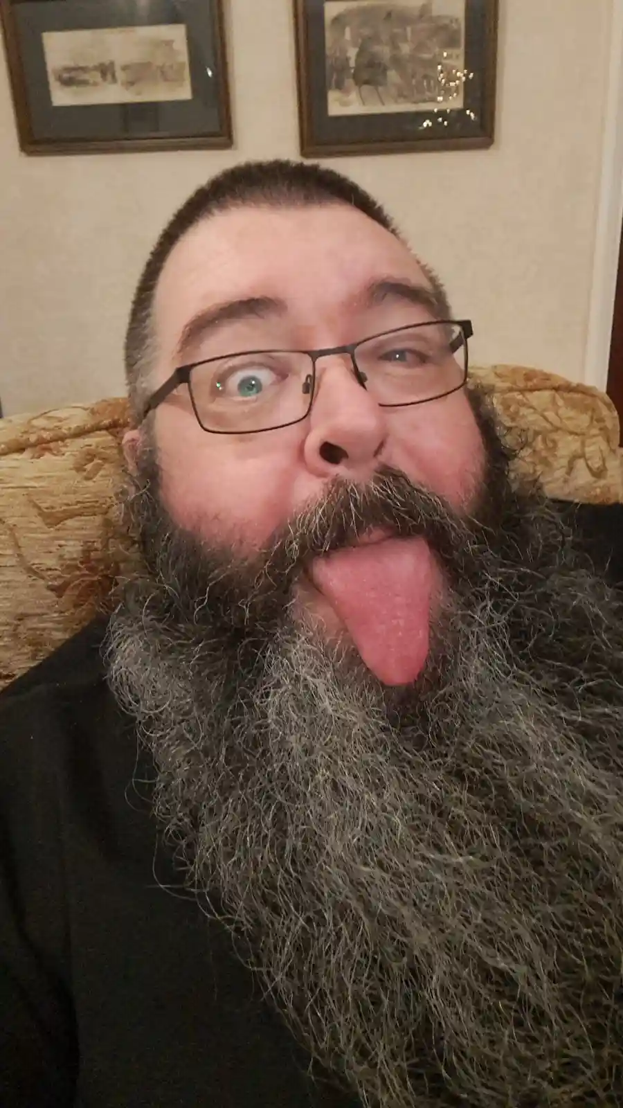 A man with a long, graying beard and glasses makes a silly face by sticking his tongue out and widening his eyes, seated on…