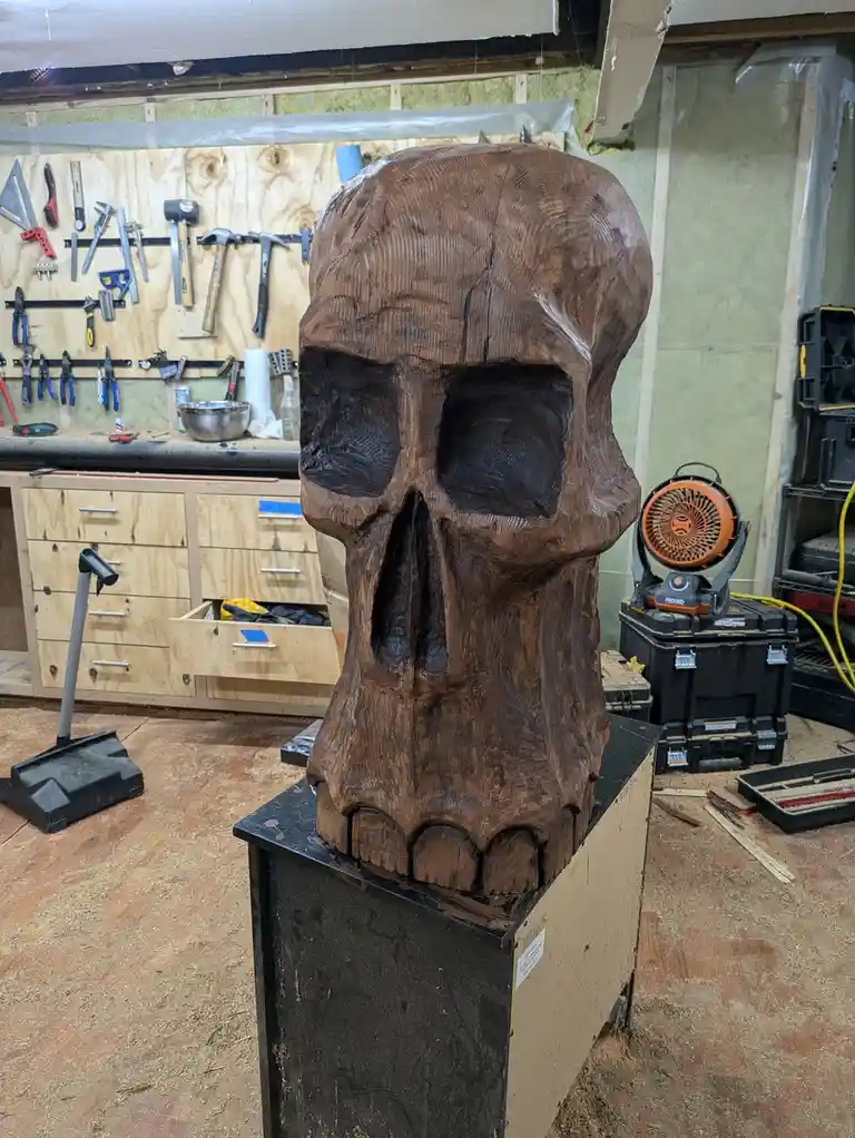 A large, intricately carved wooden skull sits on a workbench in a cluttered workshop filled with tools, sawdust, and storage…
