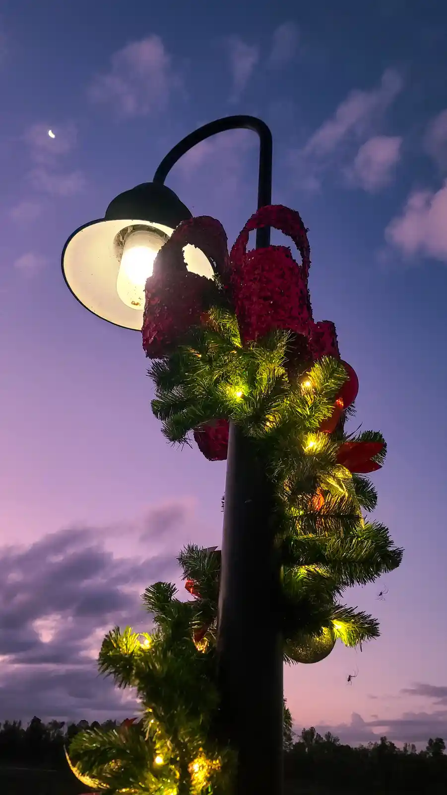 A street lamp decorated with a large red bow, green tinsel garland with yellow lights, and red ornaments, set against a…