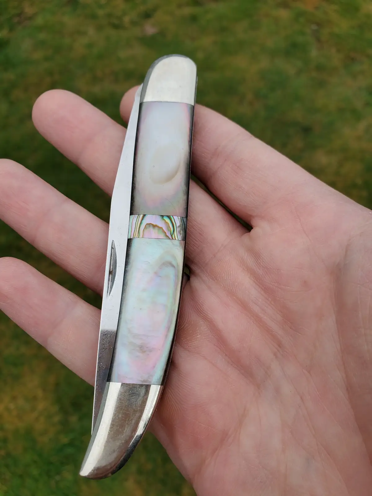 A hand holds a small, ornate knife with a blade and handle featuring iridescent mother-of-pearl inlays and silver-colored…