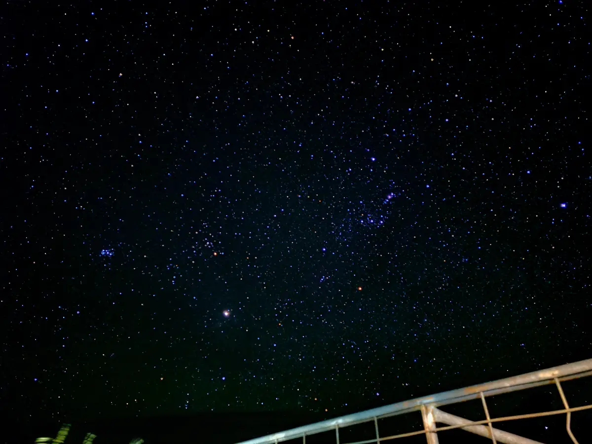 A photo of a starry night sky with countless stars visible against a dark background, framed by the top of a metal railing…