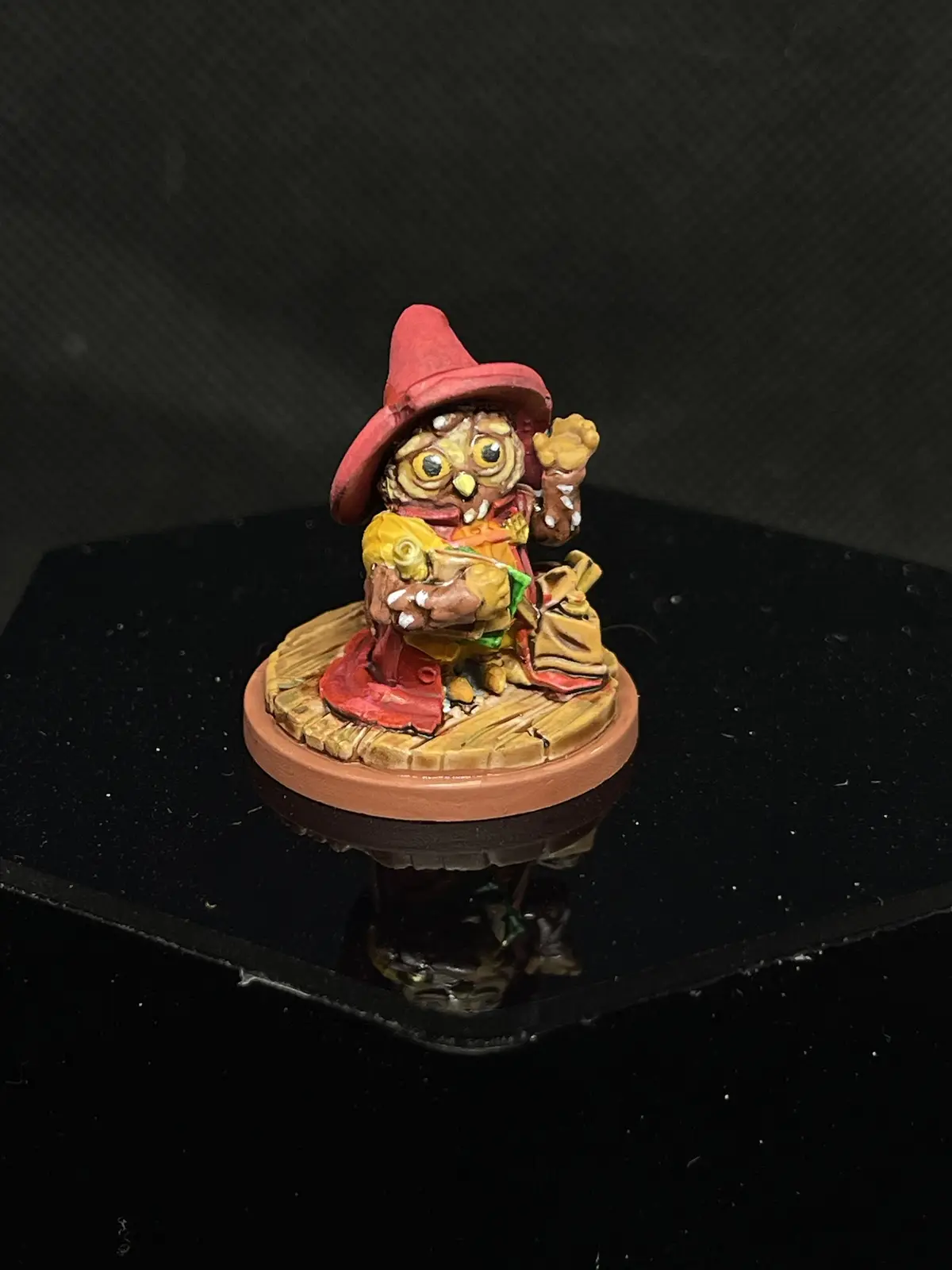 A painted miniature figurine of a small, grotesque creature with large yellow eyes, wearing a red hat and holding a yellow…