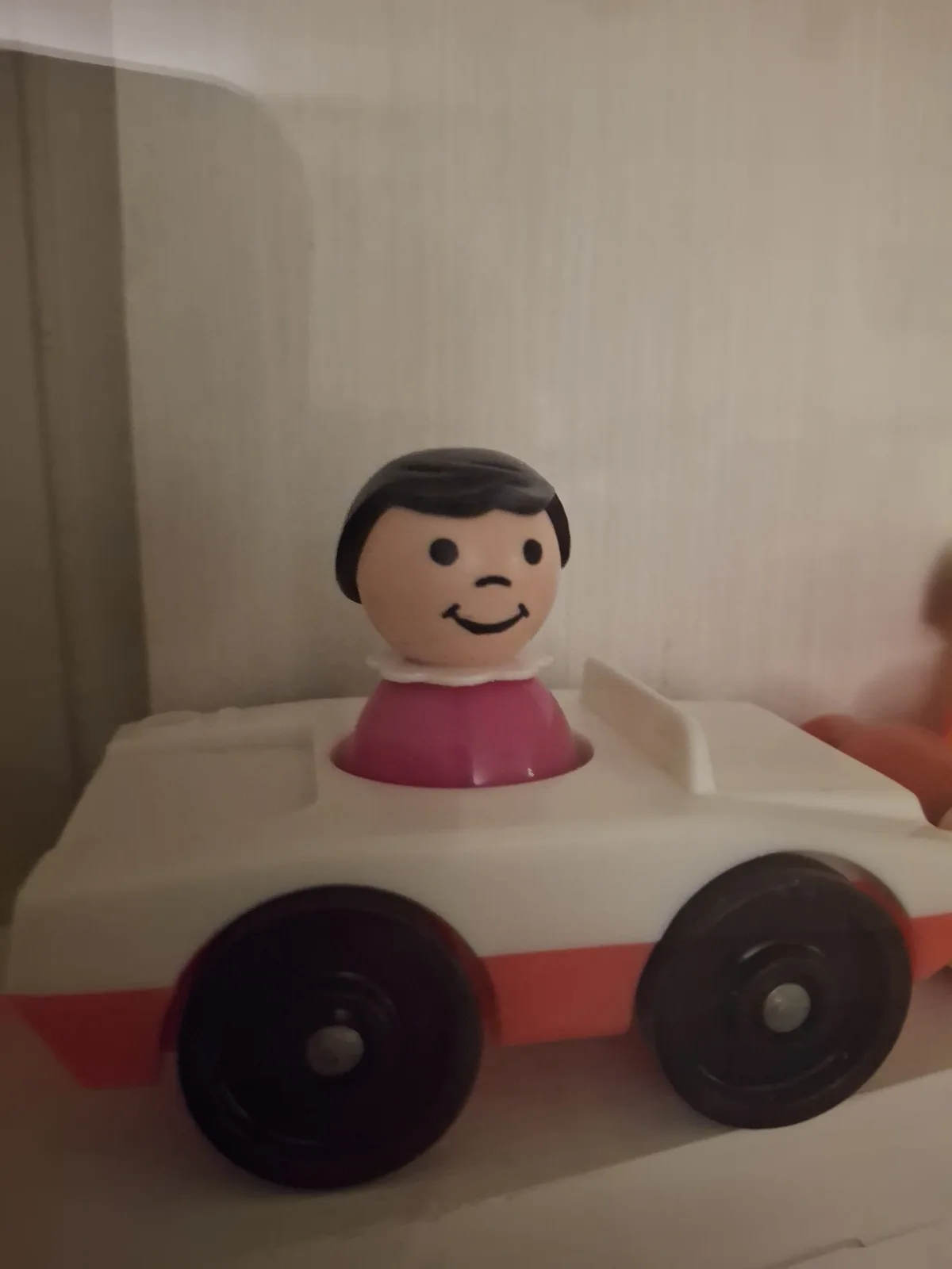 A close-up of a vintage-style plastic toy car with a smiling, cartoonish human head figure in the driver's seat. The car is…