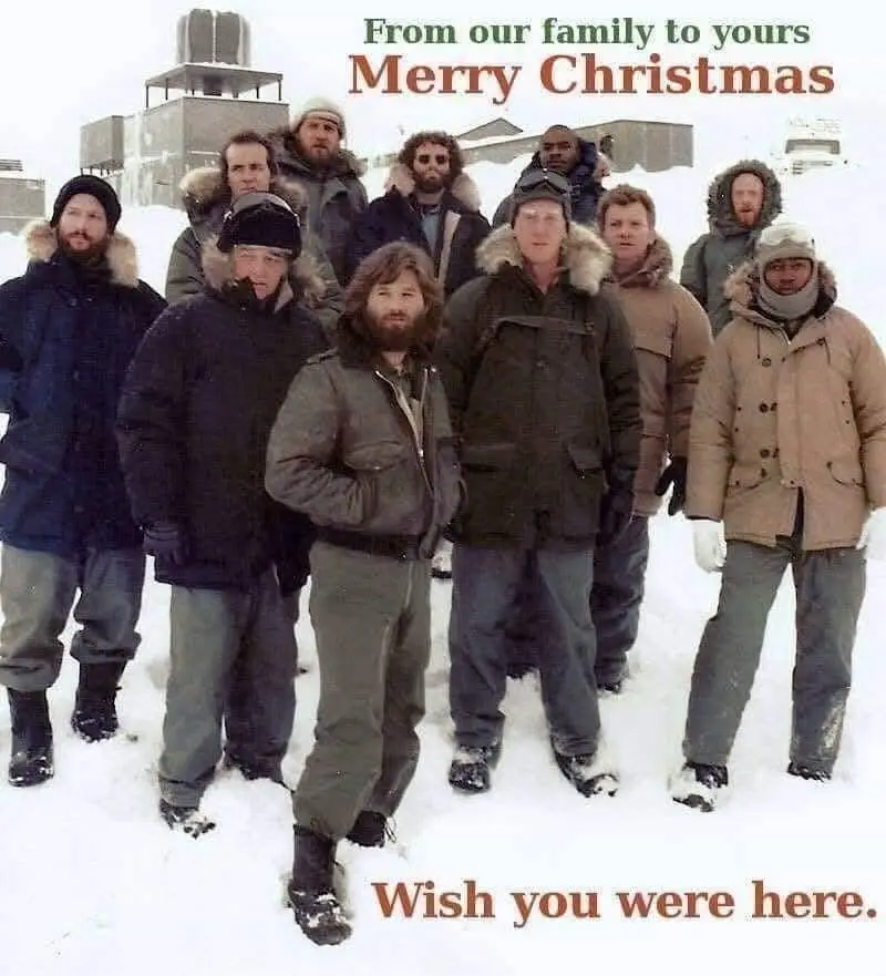 A group of men in heavy winter coats and hats stand in a snowy outdoor setting, likely a remote location, with buildings…