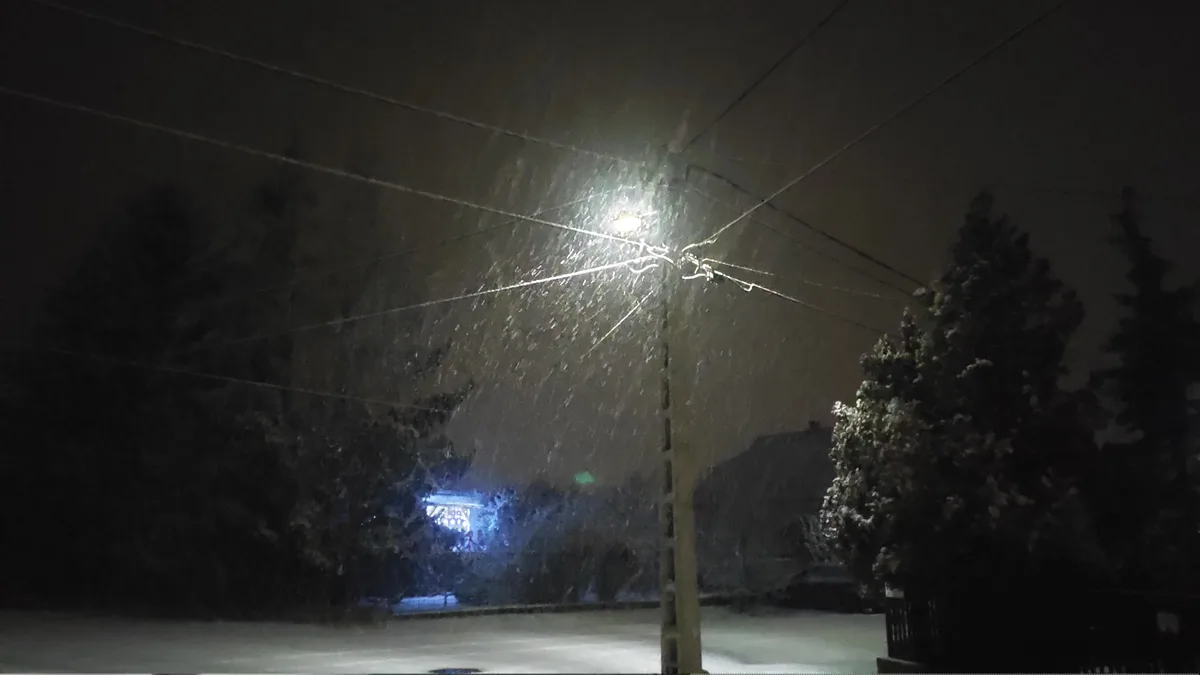 A nighttime photo taken from inside a vehicle shows heavy snow falling under a streetlight, obscuring visibility…