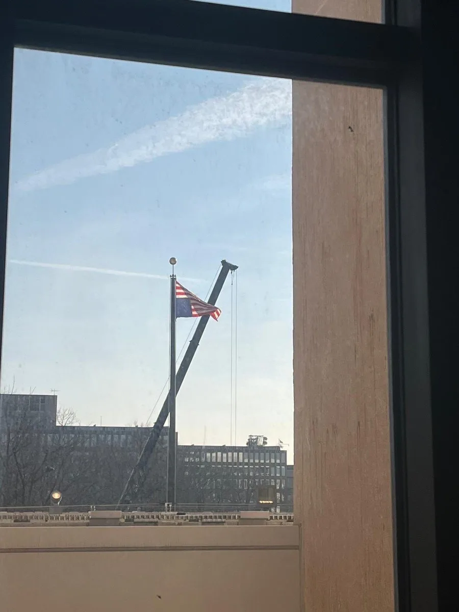A photograph taken from inside a building looking out a window shows an American flag waving on a flagpole, with a large…