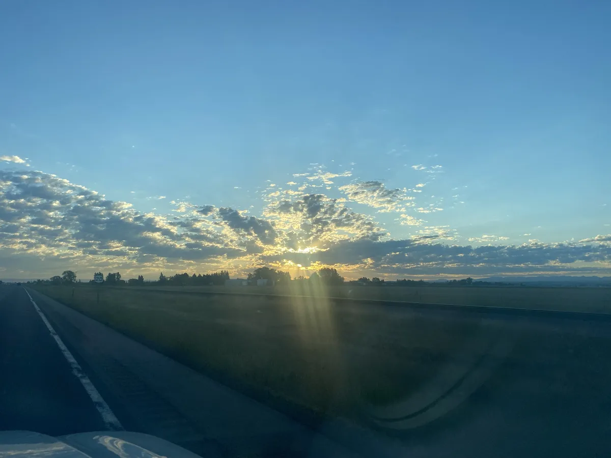 A scenic photo taken from inside a car on a highway, showing a beautiful sunrise or sunset with golden light breaking…