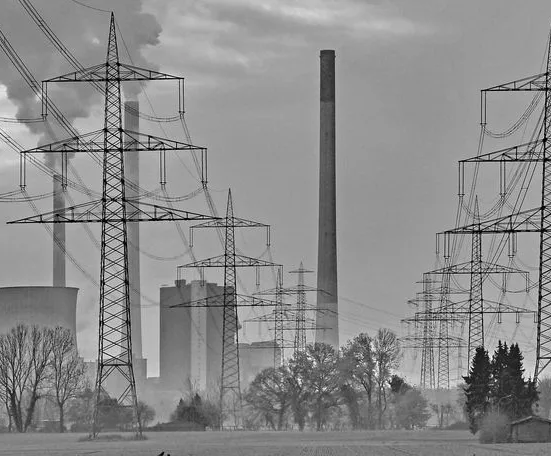 A black-and-white photograph of a large power plant with smokestacks emitting smoke, surrounded by numerous tall electricity…