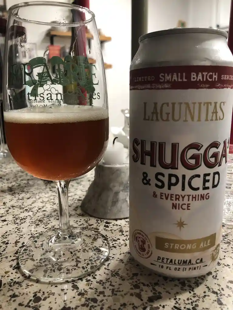 A can of Lagunitas Shugga & Spiced & Everything Nice strong ale sits next to a glass of the poured beer on a speckled…