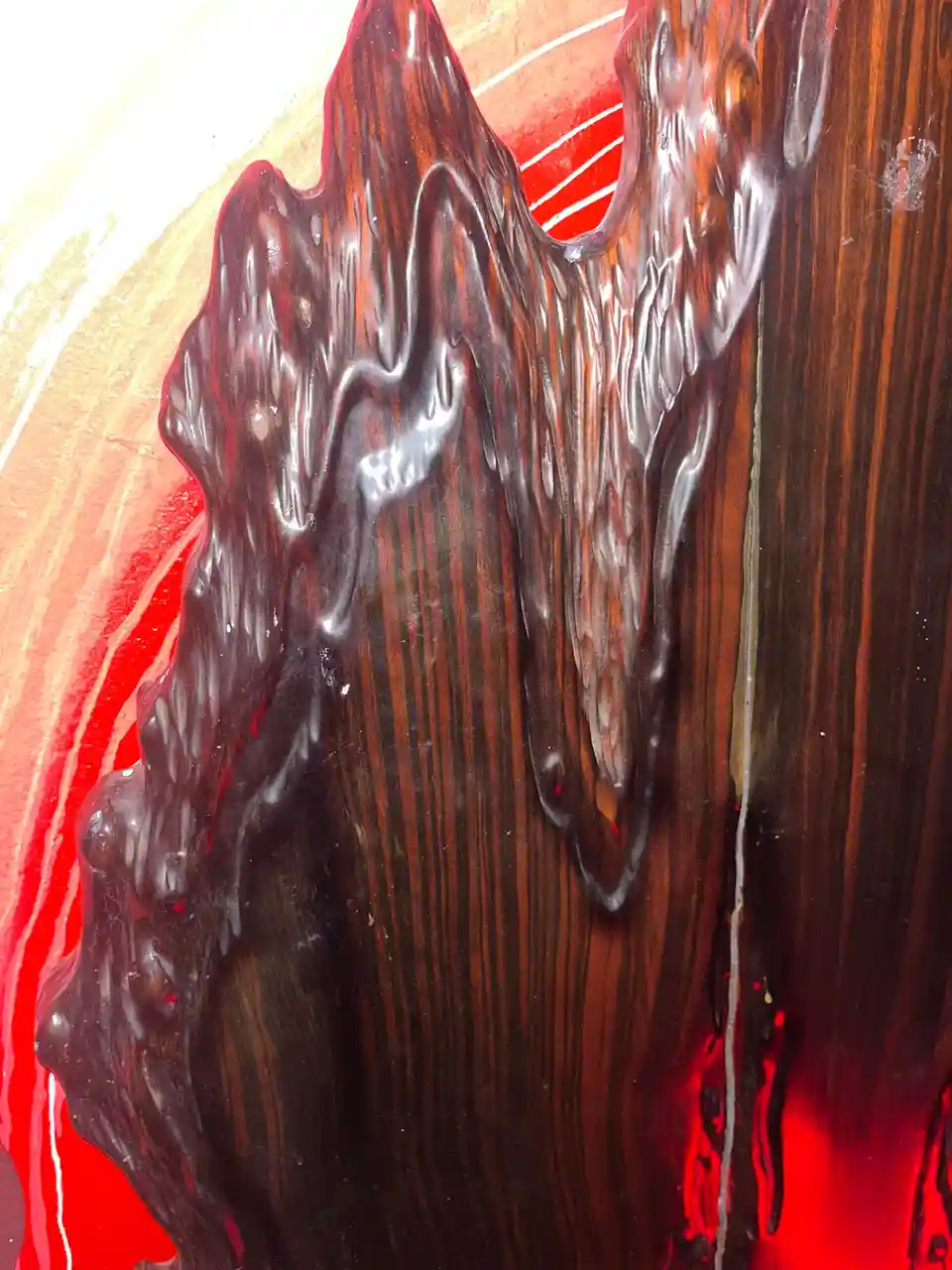 A close-up photo shows a dark, glossy, viscous substance dripping down a polished wooden surface with prominent grain, set…
