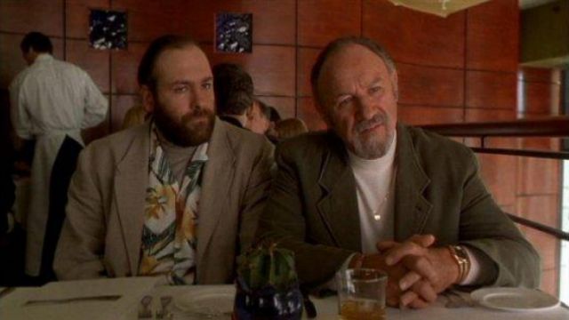 Two men sit at a restaurant table; the man on the right, with a beard and wearing a brown jacket, looks at the camera with a…