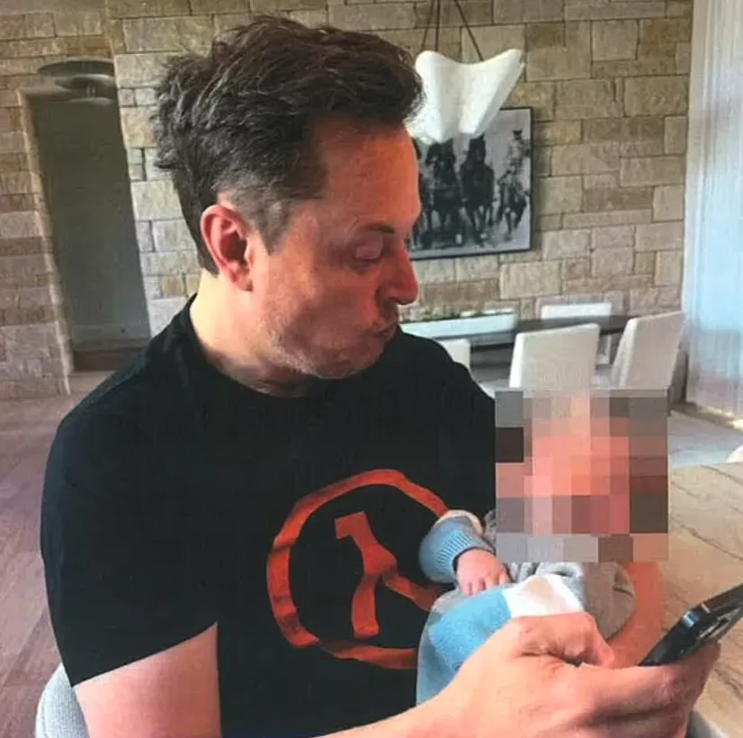 A man with dark hair wearing a black t-shirt with a red circular logo holds a baby whose face is pixelated. He is looking…