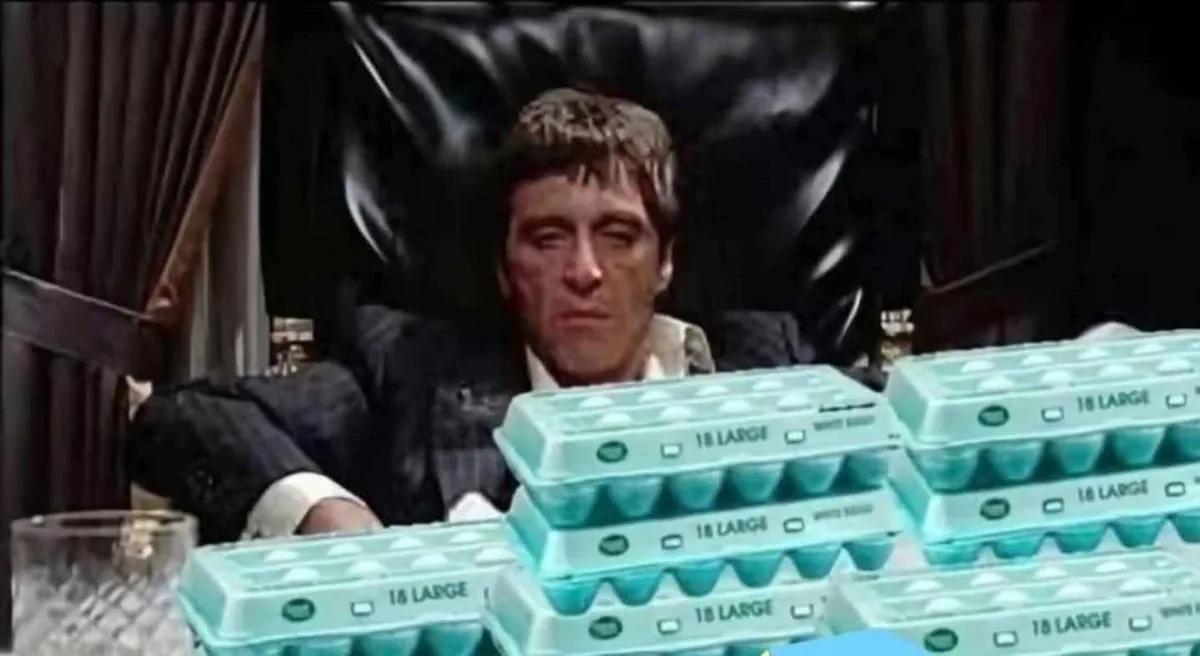 A man in a suit sits in a black leather chair behind a table piled high with multiple stacks of light blue egg cartons…