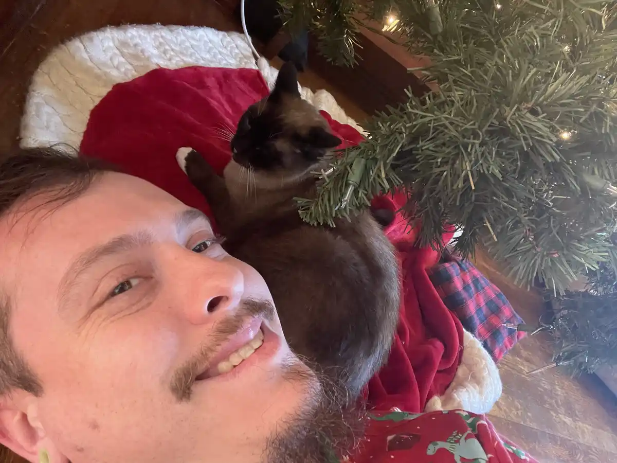 A man with a goatee and a smile lies on a red and white Santa-style blanket next to a dark brown cat with white paws. The…