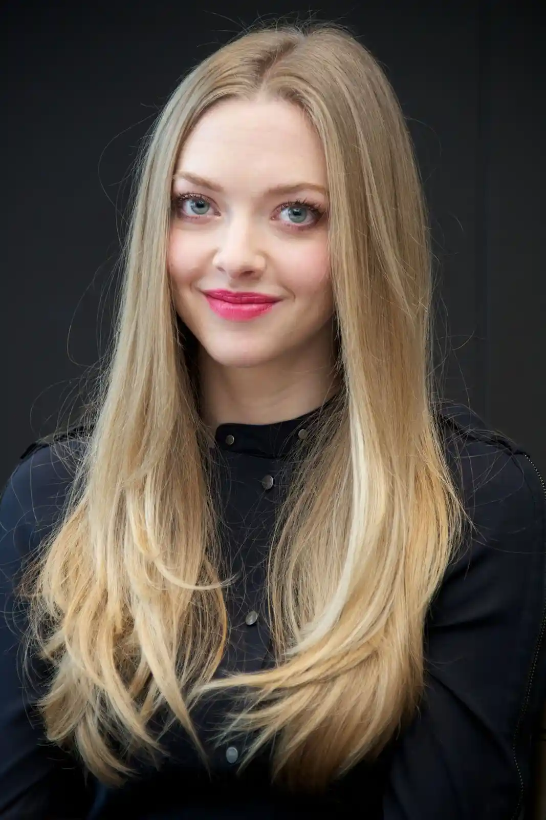 A woman with long, straight blonde hair and blue eyes smiles at the camera while wearing a black button-up shirt against a…