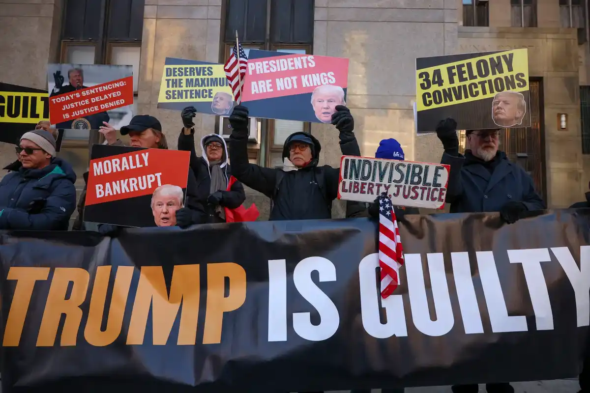A group of protesters holding signs with images of Donald Trump and slogans like '34 FELONY CONVICTIONS', 'PRESIDENTS ARE…