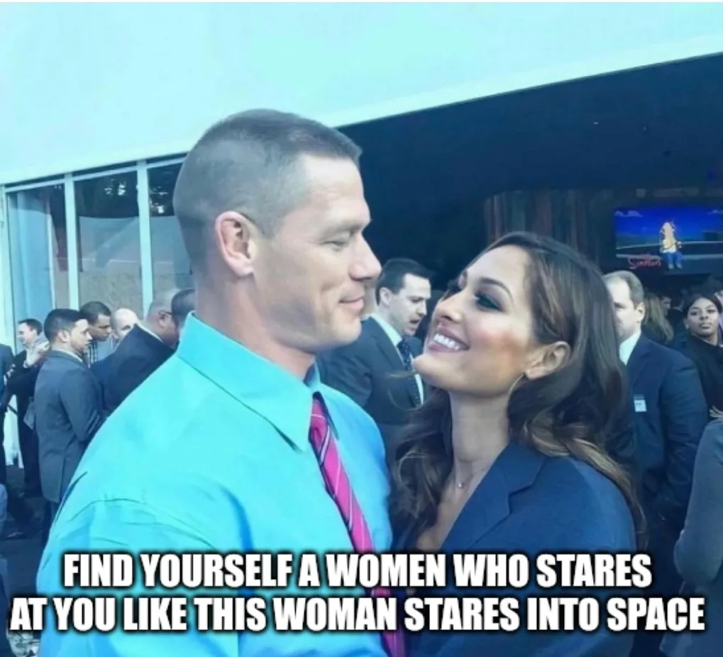 A man in a bright blue shirt and pink tie stands beside a woman in a dark blazer, both looking at each other with smiles…