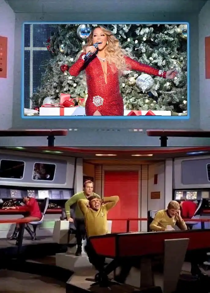 A television screen shows Mariah Carey singing in a red sequined dress in front of a decorated Christmas tree. Below, crew…