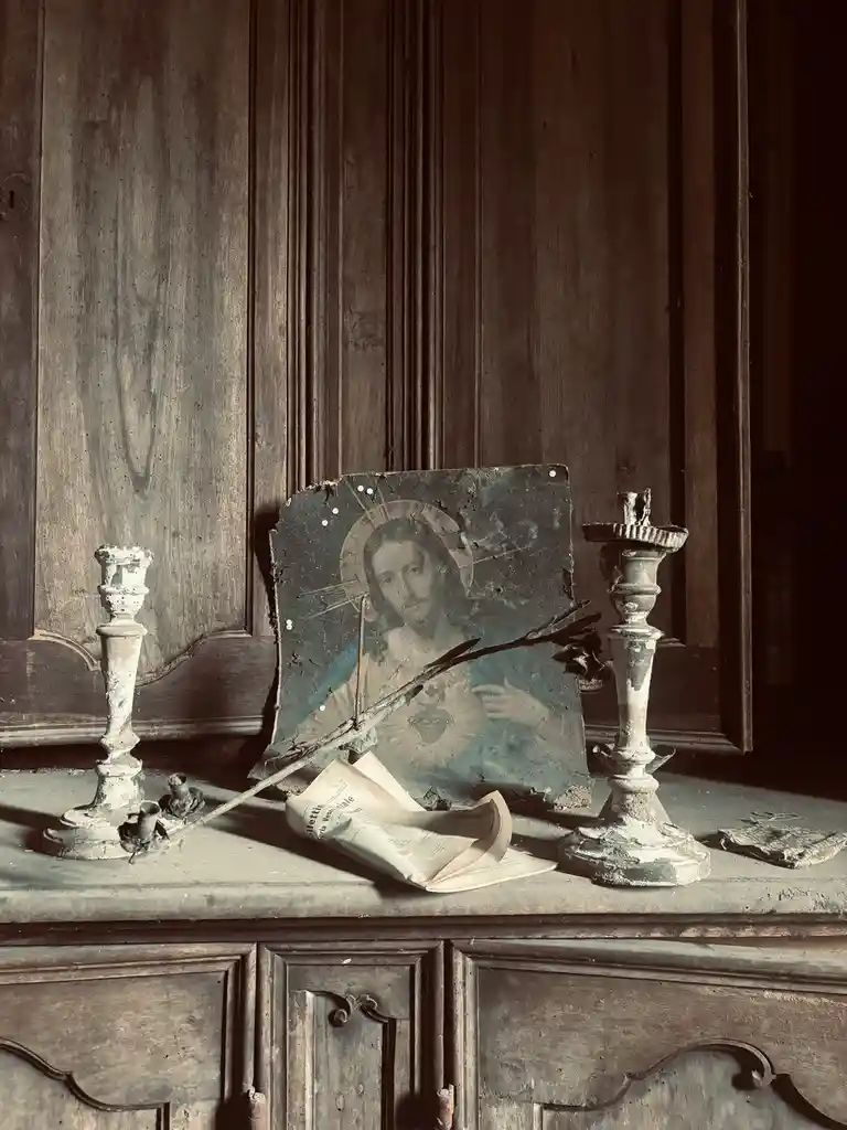 A weathered religious image of Jesus with a halo is pinned to a wooden surface, flanked by two broken candlesticks. A dried…