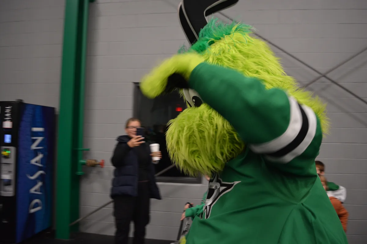 A bright green, furry mascot wearing a green jersey with a star logo is seen from behind, covering its face with one hand in…