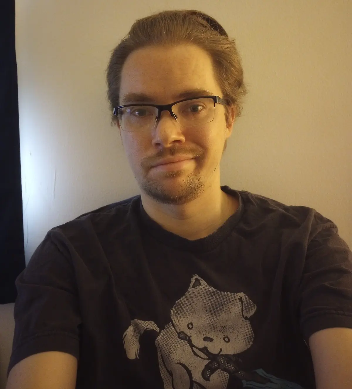A man with glasses and a goatee stares directly at the camera with a subtle, slightly smug expression while wearing a dark…