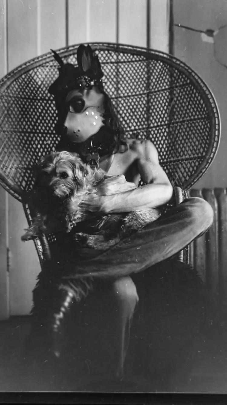 A black-and-white photo shows a person wearing a dog mask, sitting in a wicker chair and holding a small, fluffy dog. The…