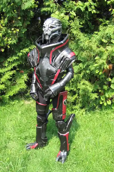 A person in a full-body, black and red armored costume with a detailed alien-like helmet stands in a grassy area with green…