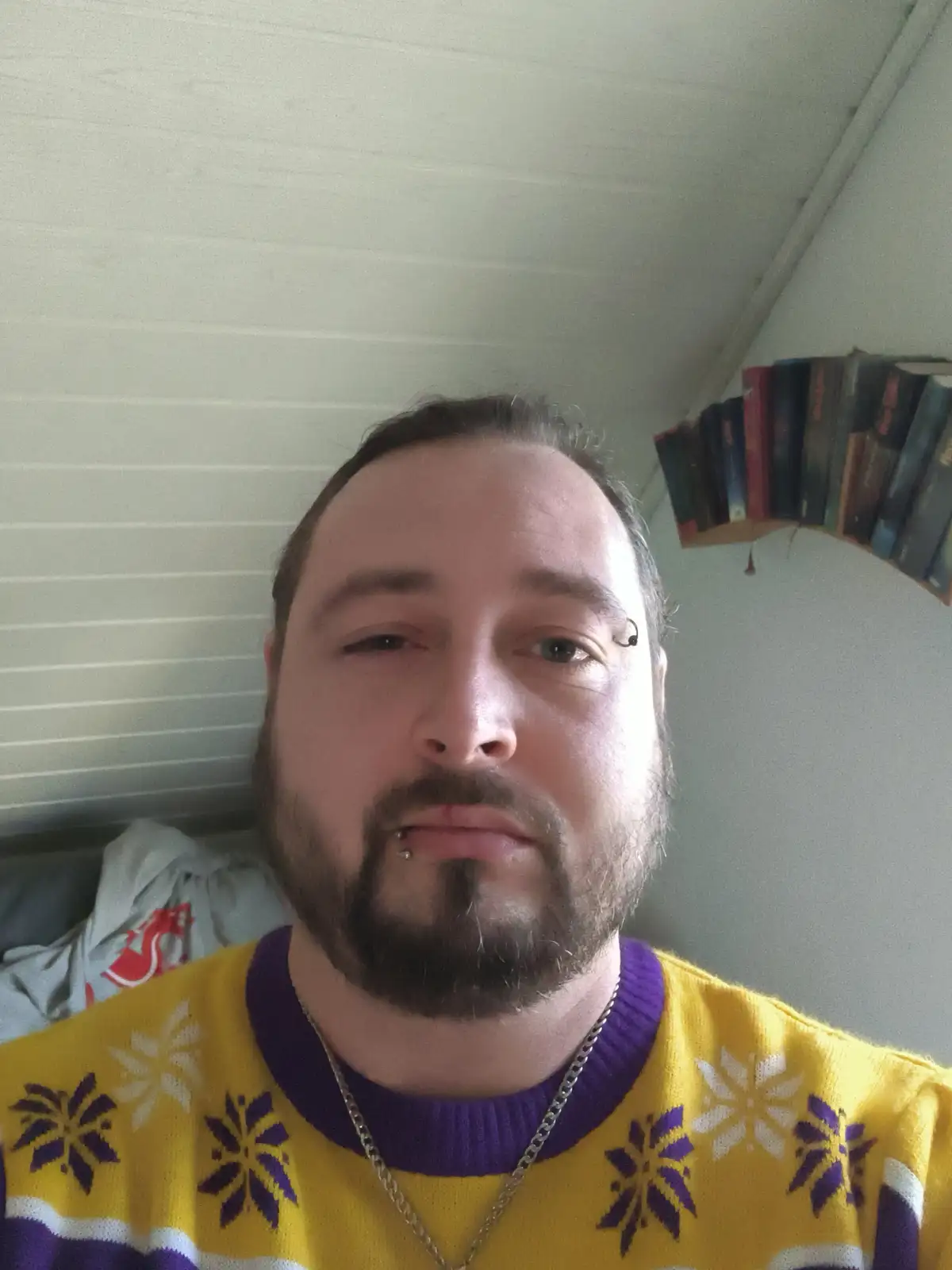 A man with a beard and multiple facial piercings stares directly at the camera with a deadpan expression while wearing a…