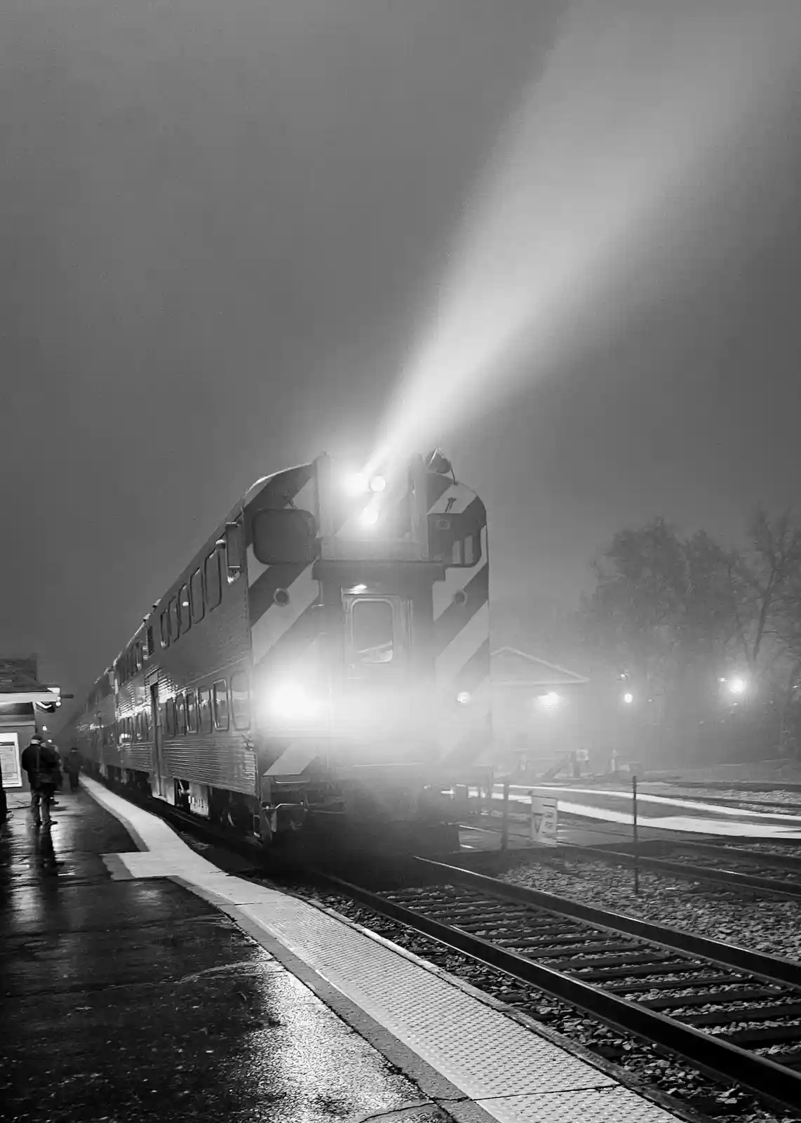 A black-and-white photo shows a train approaching a station platform at night, its bright headlights cutting through fog…