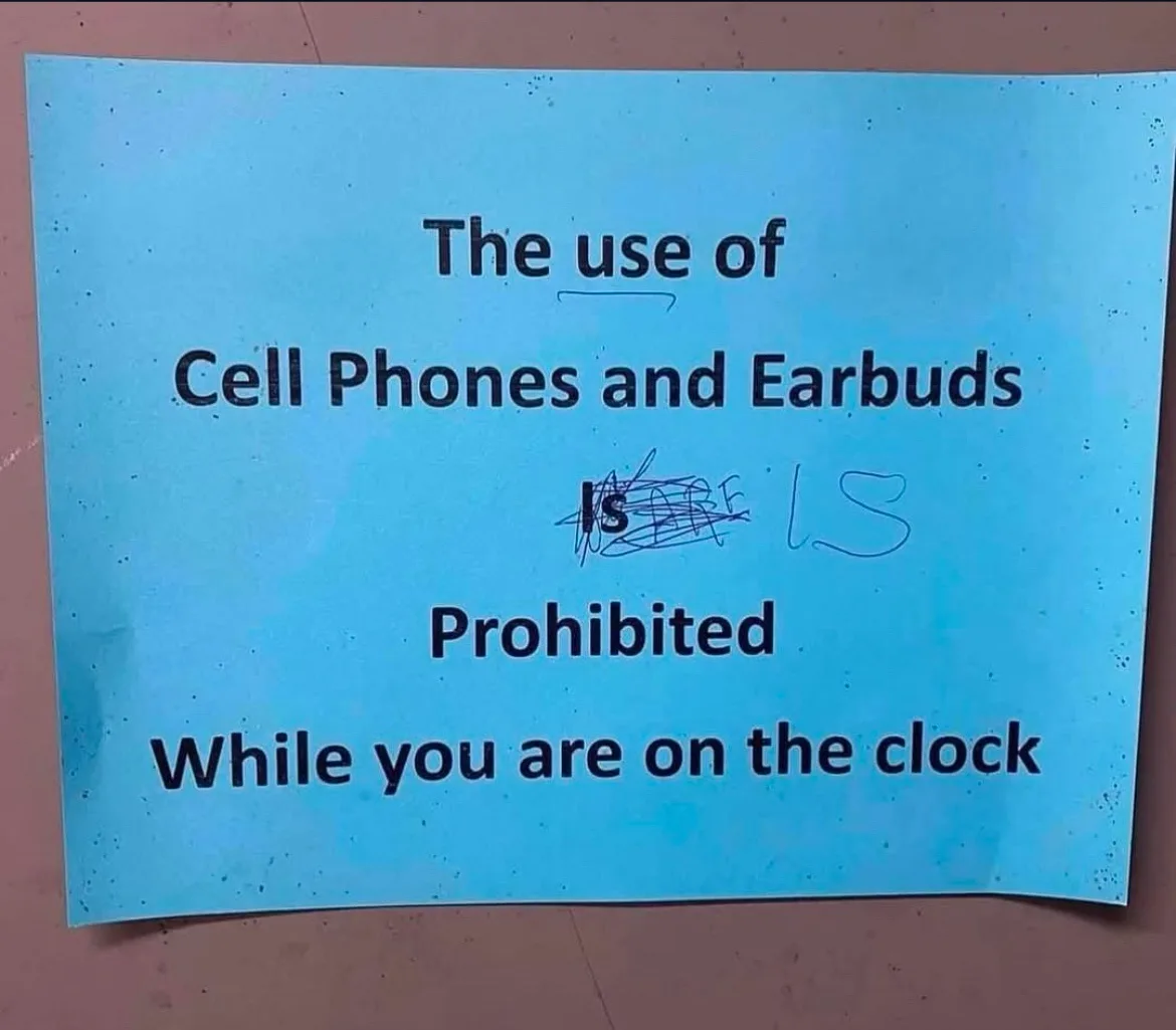 A blue sign with black text reads 'The use of Cell Phones and Earbuds IS Prohibited While you are on the clock', with the…