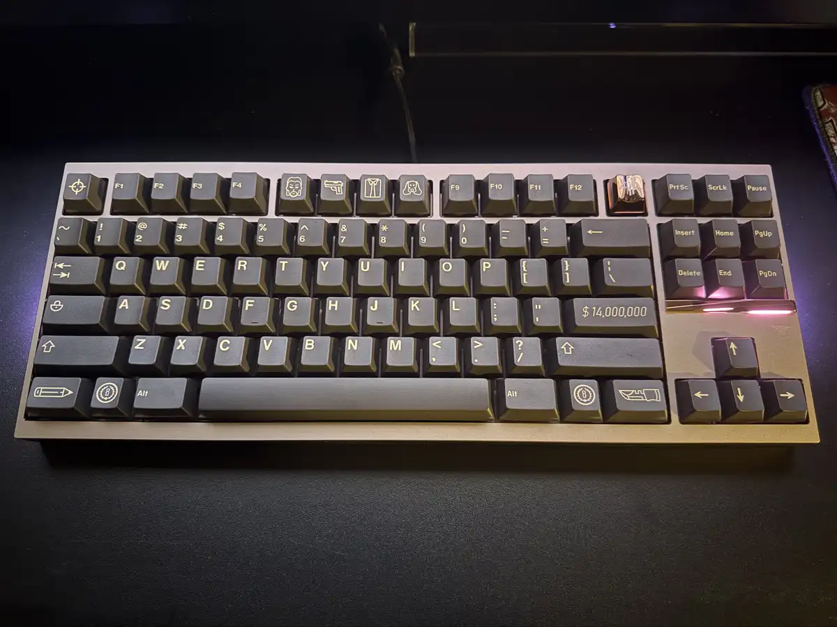 A close-up photo of a mechanical keyboard with dark grey keys and gold-colored lettering, placed on a dark surface. The…
