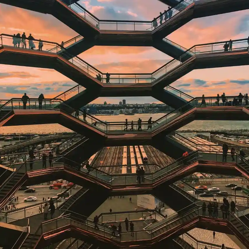 A photo of the Vessel in Hudson Yards, New York, at sunset, showing its hexagonal, multi-level structure with people walking…