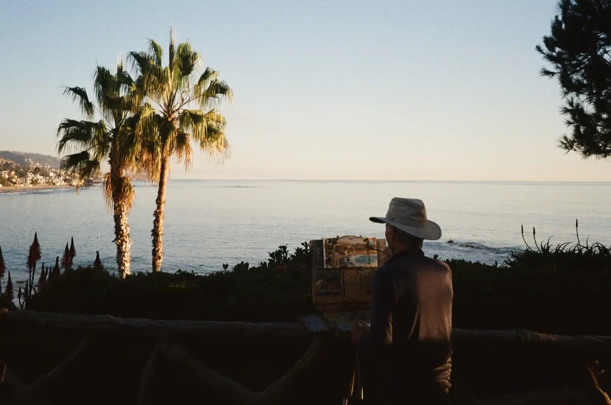 A person wearing a wide-brimmed hat is seen from behind, sitting at an easel by the ocean, painting a seascape. Palm trees…