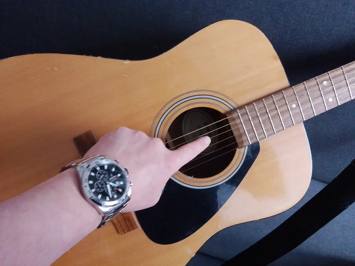 A person's hand, wearing a silver chronograph watch, points their index finger directly at the soundhole of a light-wood…