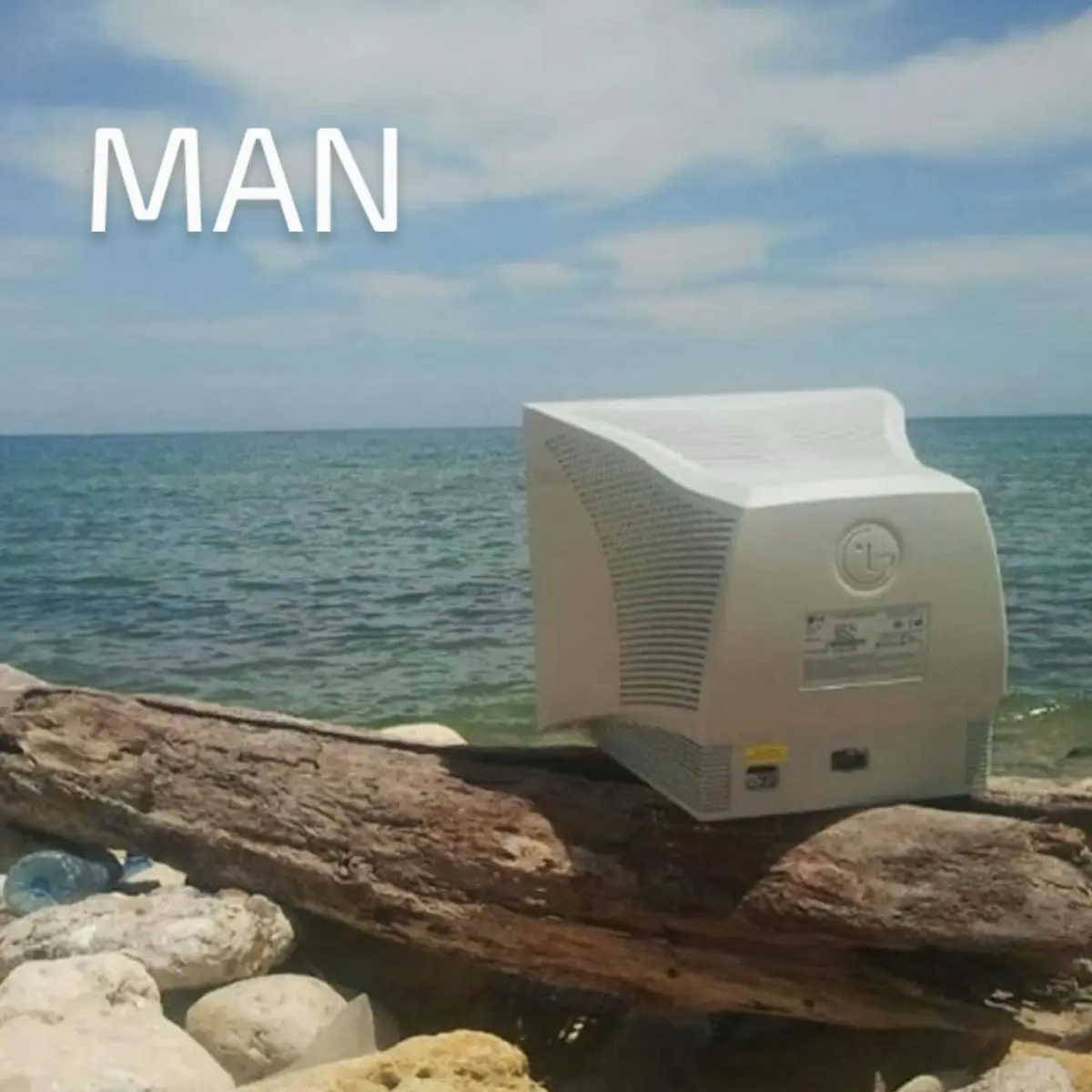 An old, white, boxy CRT computer monitor sits abandoned on a large piece of driftwood at a rocky beach, with the ocean and…