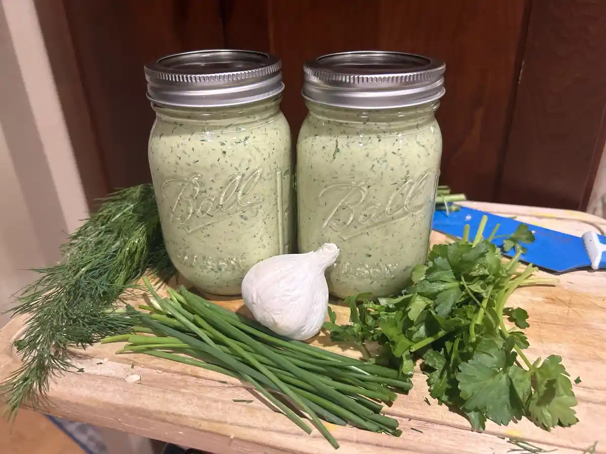 Two mason jars filled with green herb dressing sit on a wooden cutting board, surrounded by fresh dill, chives, parsley, and…