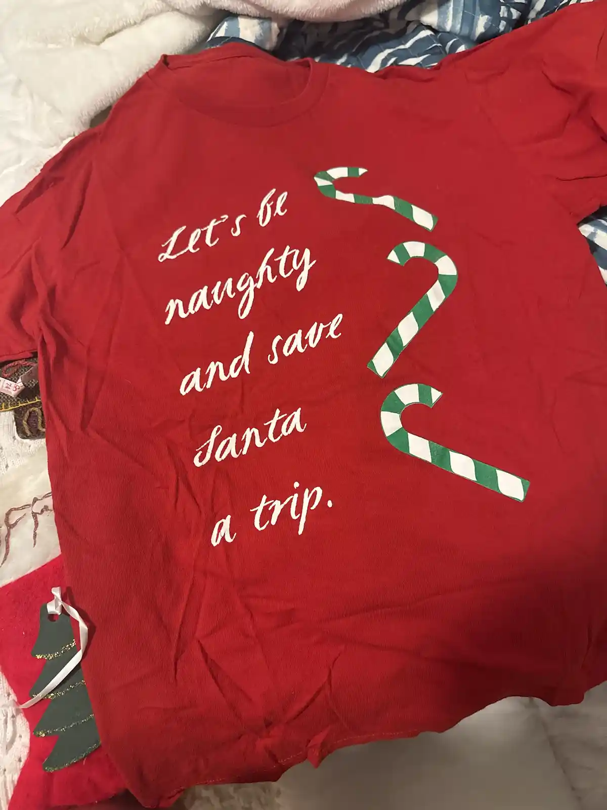 A red t-shirt laid on a bed with blankets, featuring white cursive text that reads 'Let's be naughty and save Santa a trip.'…